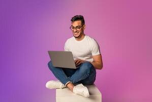 Full length of young Arab man sitting cross legged with laptop, having call, working online in neon light. Happy middle Eastern guy watching movie, chatting on internet photo