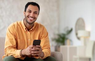 Happy Smiling Man Using Phone Browsing Internet Sitting On Couch At Home. Cheerful Cellphone User Testing New Application For Smartphone, Posing Looking At Camera, Free Copy Space, Mockup photo