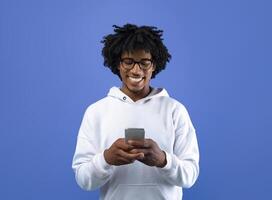 Happy black teenager looking at mobile phone screen, using new cool application on blue studio background. Joyful African American adolescent using smartphone, browsing social network photo