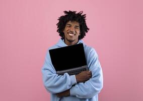 Cool African American teenager in casual wear hugging laptop pc with mockup for design on empty screen over pink studio background. Space for your website or online advertisement photo
