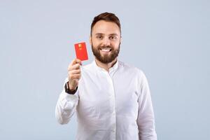 Cheerful millennial guy in formal clothes showing credit card to camera and smiling on grey studio background. Happy bank client recommending financial services, using electronic money photo