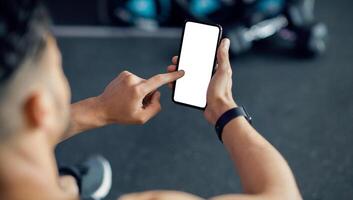 Unrecognizable Male Using Cellphone With White Screen At Gym, Mockup Image Of Sporty Man Browsing Mobile App On Blank Phone While Relaxing After Training In Sport Club, Over Shoulder View photo