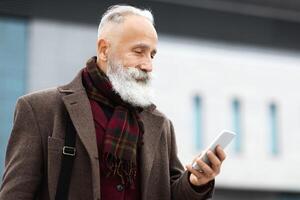 Handsome grey-haired businessman coming home from job, looking at smartphone screen, checking emails or reading messages, copy space. Wealthy senior man checking mobile phone on street photo