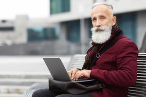Modern grandfather using laptop while sitting on bench on the street, typing on keyboard and looking at camera, writing novel or having remote job during retirement, empty space photo