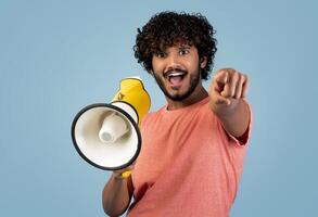 Handsome young eastern guy shouting through yellow loudspeaker and gesturing, pointing at camera, hindu man making announcement on blue studio background, copy space photo