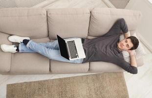 Tired guy having rest, overworked man freelancer with laptop dreaming on couch at home, top view, copy space photo