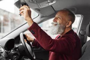 Elderly businessman in stylish suit getting ready before travelling by his auto, setting rearview mirror. Aged grey-haired man driver fixing mirror in car while driving, copy space photo