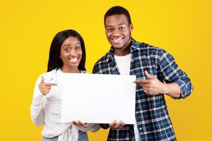 Good offer, great review and cool ad. Smiling generation z african american students guy and woman in casual hold paper banner with blank space and point fingers at it, isolated on yellow background photo