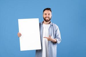 Advertising billboard. Portrait of smiling young guy pointing at blank white poster with mockup, space for your design. Happy millennial man demonstrating empty banner template photo