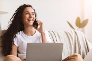 Cheerful young woman freelancer talking on mobile phone and using laptop, working from home during isolation, looking at copy space. Happy curly lady having conversation on phone photo