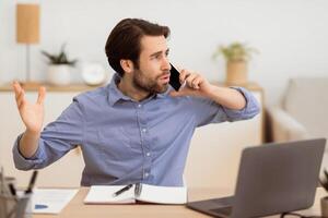 Angry Businessman Calling By Phone Having Problems Working At Laptop Sitting At Workplace In Modern Office. Aggressive Customer, Communication Issue Concept. Selective Focus photo