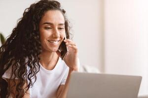 Closeup of happy young woman freelancer talking on mobile phone and using laptop, working from home during isolation, copy space. Curly lady having conversation on phone and looking at laptop screen photo