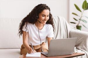 Joyful young woman with dark curly hair studying online, using laptop and headset, having webinar from home. Smiling lady sitting in front of computer, looking at screen and taking notes, empty space photo