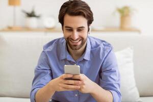 Cheerful Guy Making Call Using Smartphone And Earbuds Sitting On Sofa At Home. Man Playing Mobile Game In Living Room Indoors. Gadget, New Application Concept photo