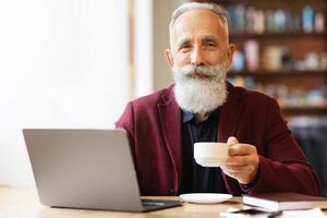 Handsome elderly man spending time at cafe, drinking coffee or tea and using laptop, websurfing or working online, smiling at camera, empty space. Active lifestyle during retirement concept photo