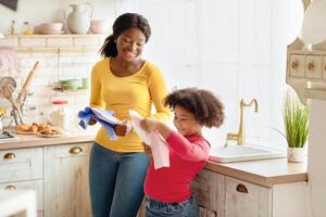 Cute African American Girl Helping Mother With Household, Wiping Dishes In Kitchen After Washing. Adorable Black Little Helper Child Enjoying Spending Time With Mom At Home, Learning Housekeeping photo