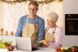 Online Grocery Shopping. Elderly Couple Using Laptop And Credit Card Buying Groceries And Ordering Food Online Standing In Modern Kitchen At Home. Happy Senior Customers Ordering Products Via Computer photo