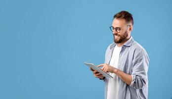 Smart millennial guy in glasses using tablet computer over blue studio background, banner design with copy space. Cheerful young man working, studying or communicating online photo