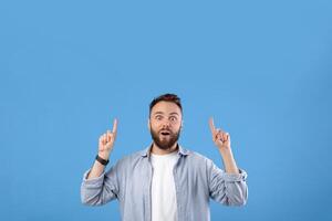 Handsome bearded guy having shocked face expression, pointing at blank space over his head on blue studio background. Millennial man offering huge discounts, promoting your product photo
