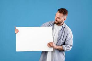 Happy smiling man displaying white blank placard on blue studio background, mockup for logo or design. Positive millennial guy with empty advertising banner, copy space photo
