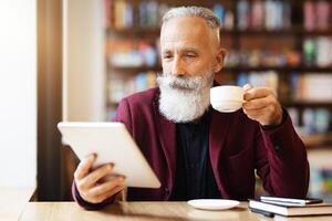 Wealthy grey-haired elderly man in burgundy jacket drinking coffee and using digital tablet, checking his email or reading book while resting at cafe, empty space. Retirement lifestyle concept photo