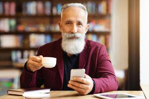 Cheerful grey-haired elderly man in burgundy jacket drinking coffee and using mobile phone, checking his email or chatting with clients, resting at cafe on his own, empty space. Retirement concept photo