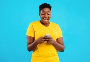 Excited African Plus-Size Woman Using Phone Application Standing Over Blue Studio Background. Black Lady Playing Mobile Game On Cellphone. Great App For Your Smartphone Concept photo