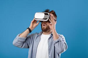 Cool millennial guy experiencing virtual reality in VR headset on blue studio background. Young bearded man using futuristic technologies, playing realistic online game, exploring cyberspace photo