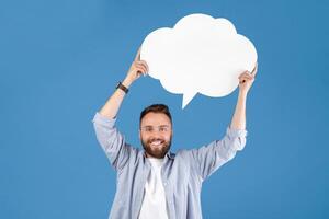 Millennial hipster guy holding speech bubble on blue studio background, mockup for design. Young Caucasian man with word cloud giving ad or presentation, sharing important info photo