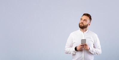 Young businessman with mobile phone deep in thought, looking aside on grey studio background. Portrait of thoughtful millennial man with modern gadget, working or studying online photo