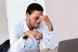 Exhausted african american manager or ceo in formal shirt suffering from burnout, sitting in front of laptop in office, rubbing eyes, looking for creative solution, lack of ideas, copy space photo