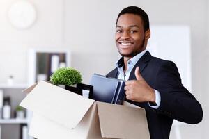 Happy black businessman moving to new office with box and his belongings, showing thumb up, copy space. Successful african american manager holding box with working stuff, changing working place photo