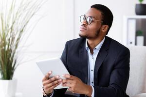 Modern technologies in business. Pensive african american young businessman in suit using digital tablet in office, working online, getting ready for presentation, looking at copy space photo