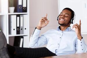 Happy young black businessman with closed eyes listening to music with legs on the table at workplace, using headphones, relaxing during difficult working day at office, free space photo