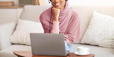 Joyful Muslim woman in hijab working online at home office, using laptop and having coffee. Smiling Arab lady having freelance job, participating in online meeting with coworkers photo