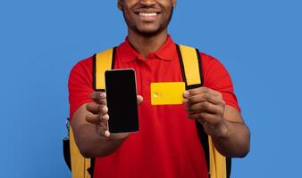 Closeup of unrecognizable smiling black delivery guy showing credit card and smartphone with blank screen for mockup, wearing red uniform and thermo backpack bag, isolated on blue background photo