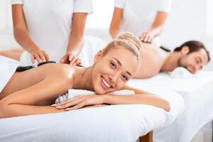 Couples Spa. Smiling Lady Getting Hot Stones Massage Enjoying Beauty Treatment With Her Husband Lying On Beds At Spa Resort Indoors. Wellness And Relaxation For Two Concept. Selective Focus photo