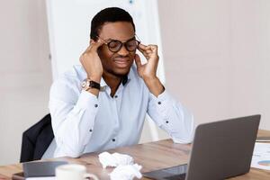 Exhausted black manager with glasses having headache while working in office in front of laptop, having financial crisis, looking for solution, copy space. Business crisis, crisis management concept photo