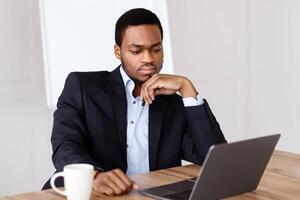 Troubled black manager looking at laptop screen, drinking coffee, having problems with his business, office interior, copy space. Crisis in business, difficuties with work concept photo