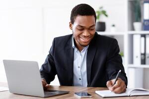 Smiling african american entrepreneur having online training in office, using laptop, taking notes, copy space. Cheerful black businessman working with computer and writing in notepad photo