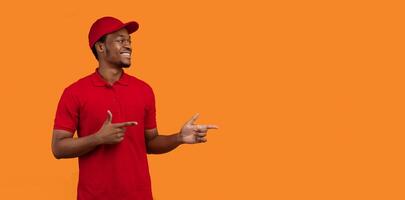 Look Here. Portrait of smiling black delivery man in red t-shirt and cap smiling, looking and and pointing fingers aside at copy space for text or logo isolated over orange studio background, panorama photo