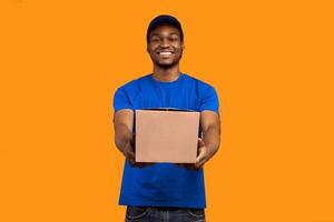 Best Service Concept. Smiling black delivery man wearing blue cap and unform holding cardboard box, showing and giving it to camera standing isolated over yellow orange studio background photo