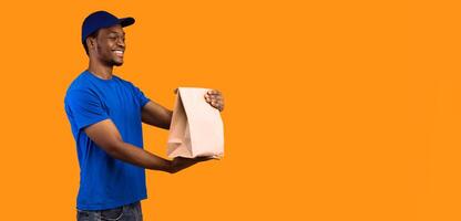 Fast Delivery. Side view of smiling african american delivery guy in blue cap holding and giving craft paper bag from restaurant with products to the side with copy space, orange background photo