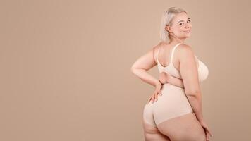 Body Positive. Beautiful Curvy Woman Posing In Underwear Over Beige Studio Background, Attractive Plus Size Blonde Female In Lingery Standing Half-Turned And Smiling At Camera, Copy Space photo