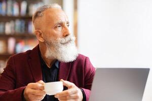 Rich senior man in stylish suit drinking coffee at cafe, sitting in front of modern laptop and looking at copy space. Elderly man using wi-fi while enjoying herbal tea, websurfing or looking for job photo