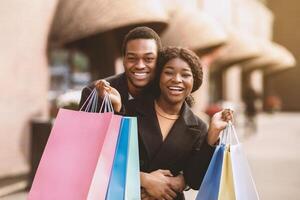 Great date and big seasonal sale during Black Friday in autumn. Young attractive happy african american guy and woman hugging, holding many colored shopping bags and look at camera near store, outdoor photo