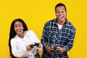 Online games together and entertainment at home. Joyful generation z african american students male and lady playing with gamepads, isolated on yellow background, empty space, studio shot photo