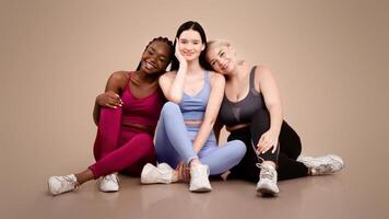 Multi ethnic female friends in sportswear posing in studio, diverse happy women of different size and appearance sitting on floor over beige background and smiling at camera, copy space photo