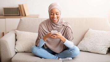 Charming black lady in hijab messaging on smartphone while sitting on cozy sofa at home. Pretty Muslim woman texting to friend, checking email, watching on mobile device indoors photo
