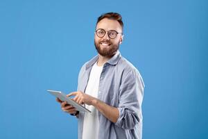 Positive young guy in casual wear using tablet computer over blue studio background. Happy millennial man with touch pad working or studying online, sending email, watching photo